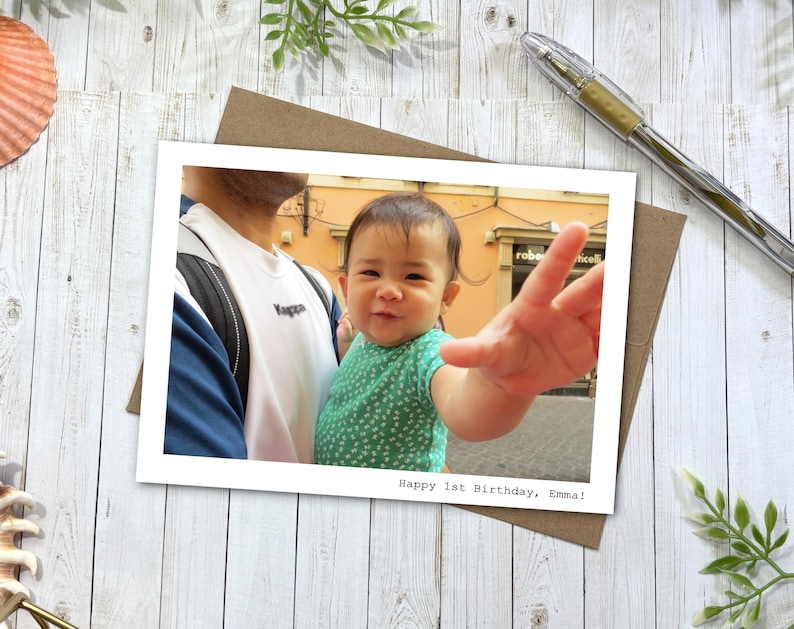 May include: A rectangular photo print featuring a baby in a green patterned shirt reaching toward the camera, held by a person in a white and blue shirt. The bottom right corner of the white border contains the text, Happy 1st Birthday, Emma!