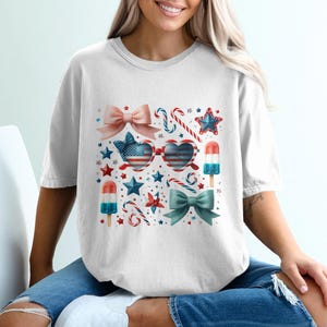 May include: White t-shirt featuring a patriotic design with bows, stars, candy canes, and heart-shaped sunglasses with an American flag pattern. The design includes red, white, and blue colors, and popsicle illustrations.