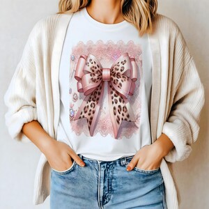 May include: White t-shirt featuring a large pink bow with a leopard print design. The bow is set against a pink lace background with decorative jewels. The shirt is worn by a person wearing a cream-colored cardigan and blue jeans.