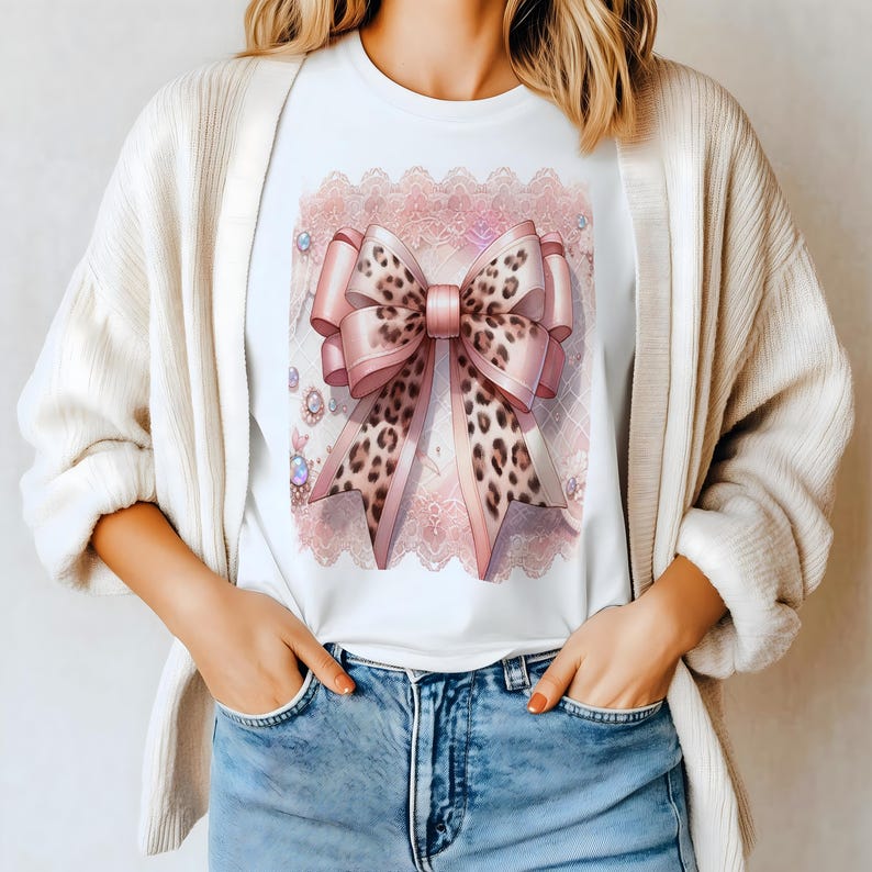 May include: White t-shirt featuring a large pink bow with a leopard print design. The bow is set against a pink lace background with decorative jewels. The shirt is worn by a person wearing a cream-colored cardigan and blue jeans.