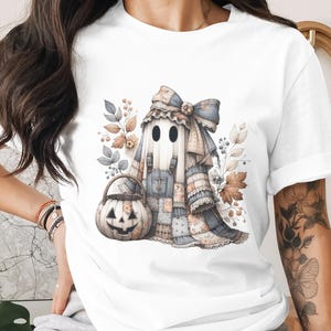 Könnte beinhalten: Weißes T-Shirt mit einer Aquarellillustration eines Geistes in einem Patchwork-Outfit, einer großen Schleife und einem Kürbis-Eimer. Das Design umfasst Blumen- und Blätterakzente, ideal für Halloween.