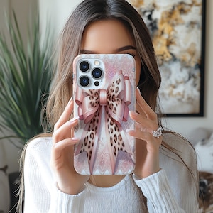 May include: A phone case featuring a large pink bow with a leopard print center. The case has a light pink background with a lace pattern. The phone case is held in front of a person's face.