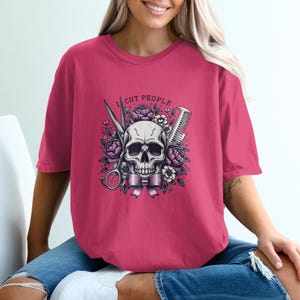 Pode incluir: T-shirt magenta com um desenho de caveira, tesoura, pente e elementos florais. O texto "I CUT PEOPLE" está acima da caveira. A t-shirt é de manga curta e gola redonda.