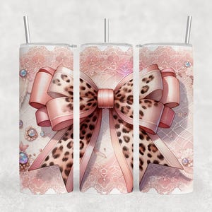 May include: A set of three stainless steel tumblers with a pink bow design. The bow features a leopard print center and pink ribbons. The background has a lace pattern with decorative jewels and a silver straw.