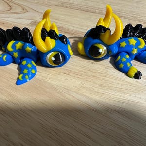 May include: Two blue, yellow, and black fantasy creatures with large eyes and star and crescent moon patterns. The creatures have yellow horns and black spiky backs. They are lying on a wooden surface.