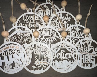 May include: A collection of white wooden Christmas ornaments with various religious phrases and imagery. Each ornament is circular, laser-cut with text and designs, and has a twine hanger with a wooden bead. The phrases include "O Come Let Us Adore Him" and "Christmas Begins With Christ."