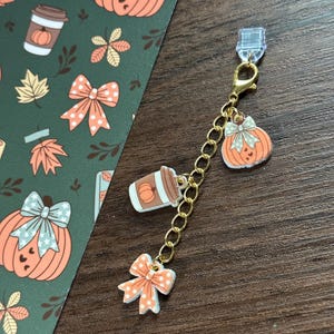 May include: A gold-toned metal chain charm featuring three acrylic pendants: an orange pumpkin with a light blue bow, a brown coffee cup with a pumpkin graphic, and an orange bow with white polka dots, attached to a clear plastic clip.