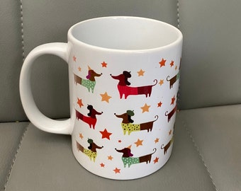 May include: White ceramic mug featuring a repeating pattern of dachshund dogs in various colors and outfits, interspersed with orange stars. The dogs have different colored bodies, hats, and outfits. The mug has a white handle.
