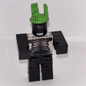 May include: A black and green toy figure with a skeleton torso and a green helmet. The figure has a black cylindrical head with red eyes and a green helmet with holes. The figure is made of plastic.