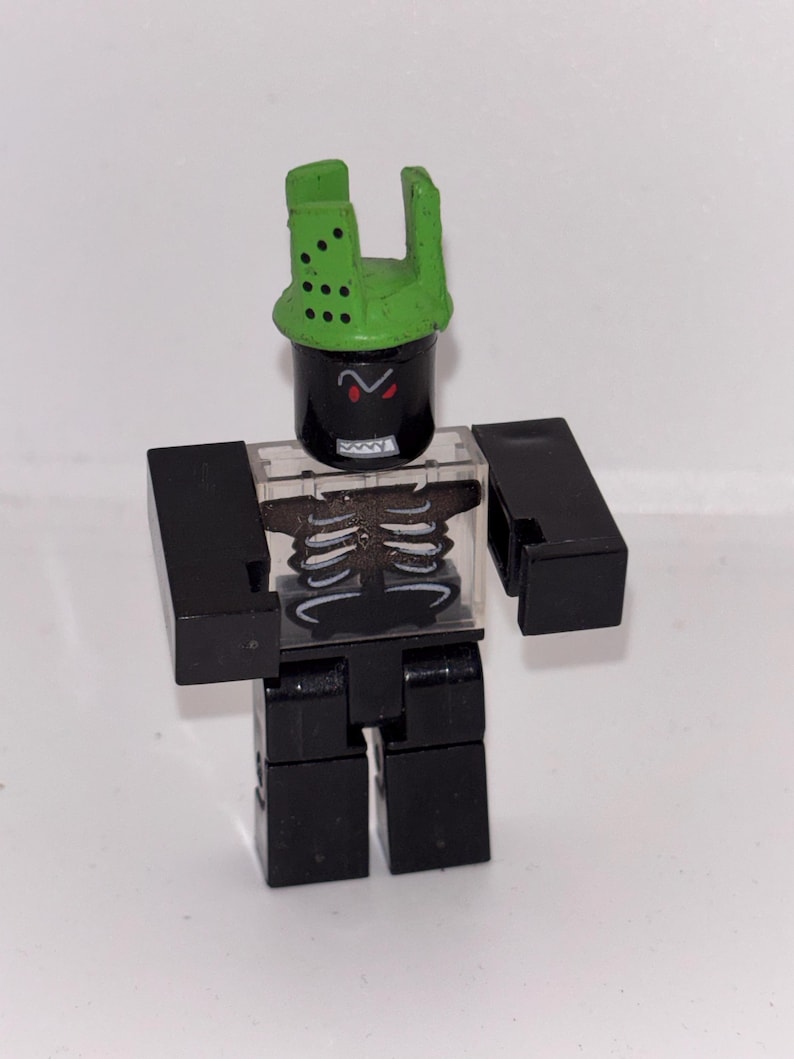 May include: A black and green toy figure with a skeleton torso and a green helmet. The figure has a black cylindrical head with red eyes and a green helmet with holes. The figure is made of plastic.
