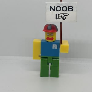May include: A Roblox-themed figurine with a red cap and a blue shirt, holding a sign that says "NOOB" with a pointing hand. The figure has yellow arms and head, green legs, and the letter "R" on the shirt and cap.
