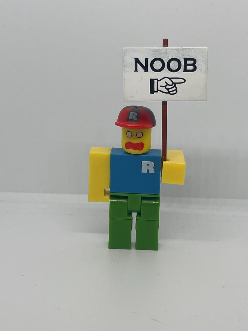 May include: A Roblox-themed figurine with a red cap and a blue shirt, holding a sign that says "NOOB" with a pointing hand. The figure has yellow arms and head, green legs, and the letter "R" on the shirt and cap.