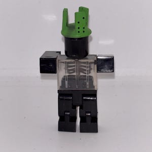 May include: A black and clear plastic toy figure with a green headpiece. The figure has a black cylindrical head, clear torso with a skeleton design, black arms, and black legs. The figure is standing against a white background.
