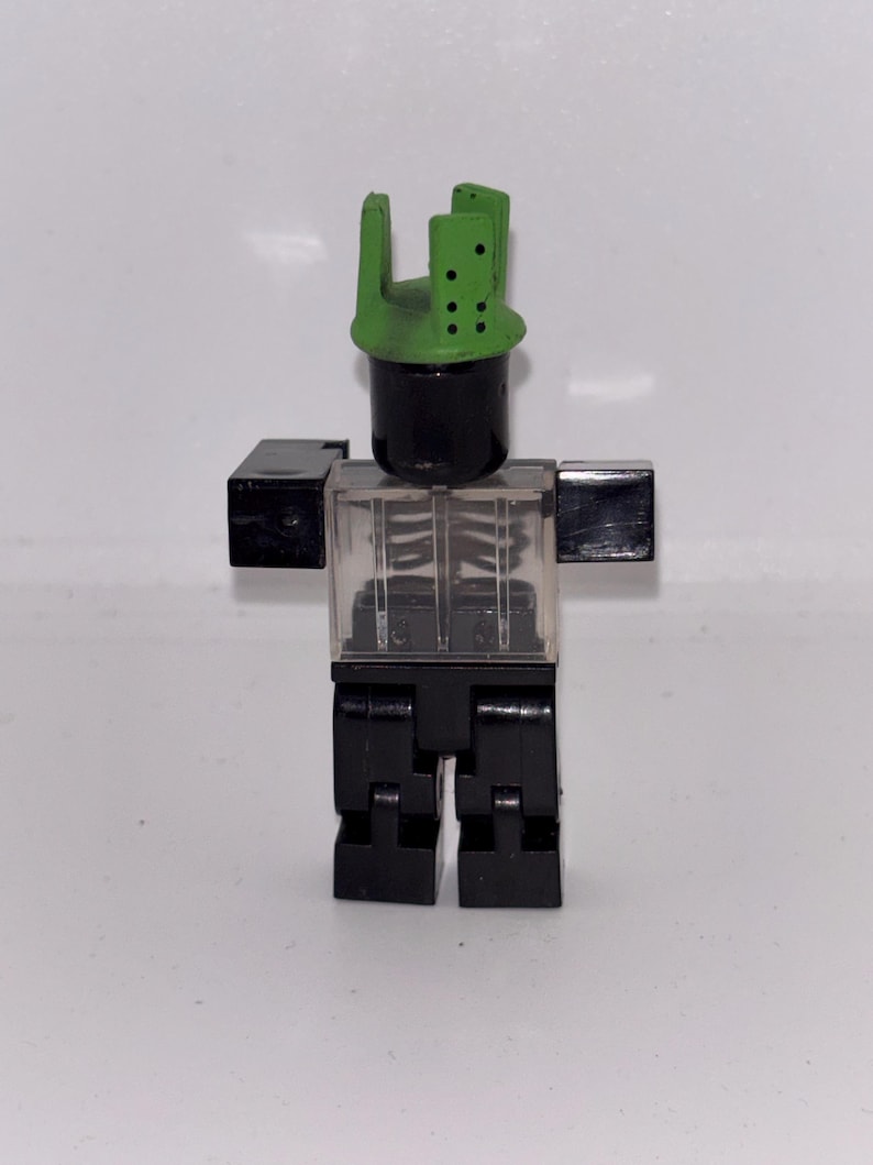 May include: A black and clear plastic toy figure with a green headpiece. The figure has a black cylindrical head, clear torso with a skeleton design, black arms, and black legs. The figure is standing against a white background.