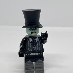 Puede incluir: Una minifigura de Lego de un personaje con traje negro, sombrero de copa y pantalón gris. La figura tiene una cabeza verde pálido con ojos enojados y boca amarilla. El torso presenta un diseño de chaleco con un símbolo de murciélago en la cintura.