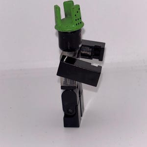 May include: A black and green toy figure with a green helmet-like headpiece. The figure is constructed from interlocking blocks, with a cylindrical head and rectangular arms and legs. The figure is posed in a standing position.