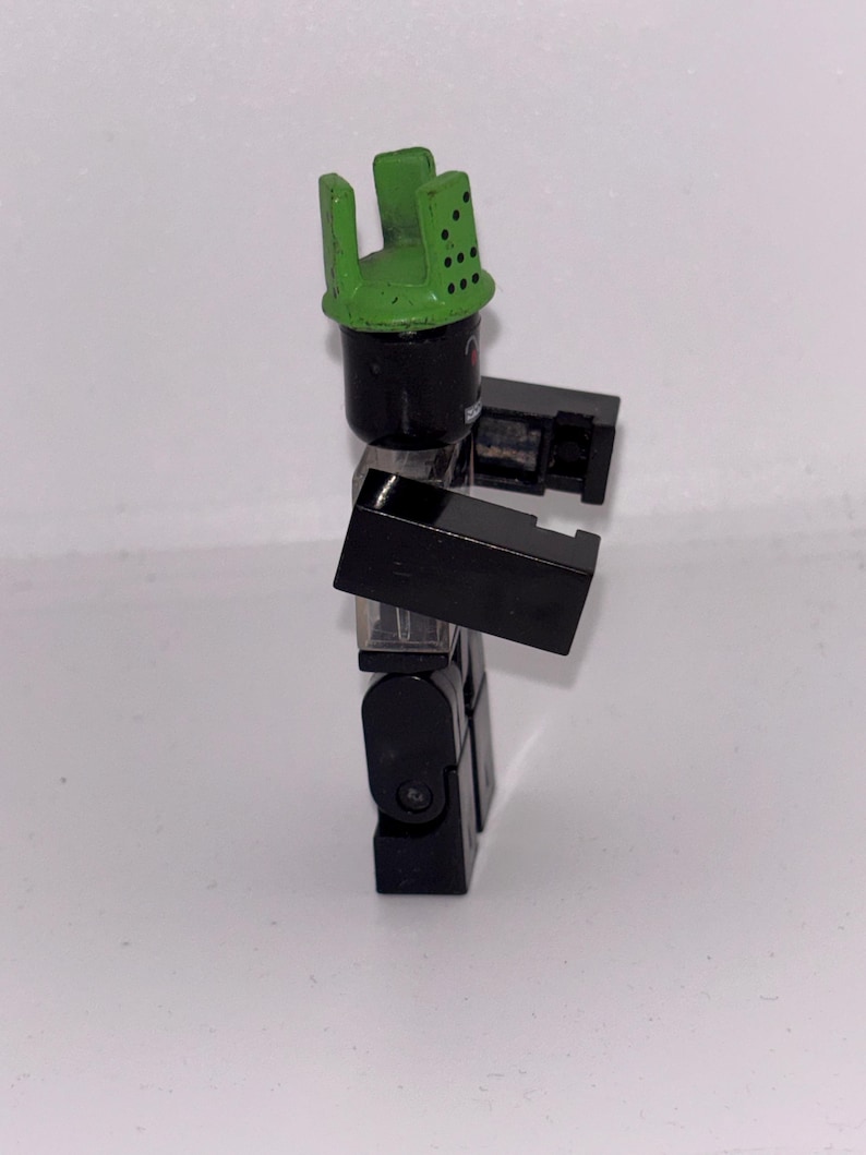 May include: A black and green toy figure with a green helmet-like headpiece. The figure is constructed from interlocking blocks, with a cylindrical head and rectangular arms and legs. The figure is posed in a standing position.