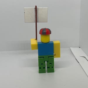 May include: A Roblox-themed figurine with a yellow head, blue shirt, green pants, and yellow arms. The figurine holds a blank white sign on a brown stick. The figure wears a red and black hat.
