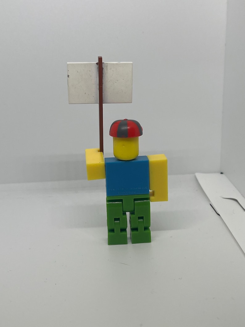 May include: A Roblox-themed figurine with a yellow head, blue shirt, green pants, and yellow arms. The figurine holds a blank white sign on a brown stick. The figure wears a red and black hat.