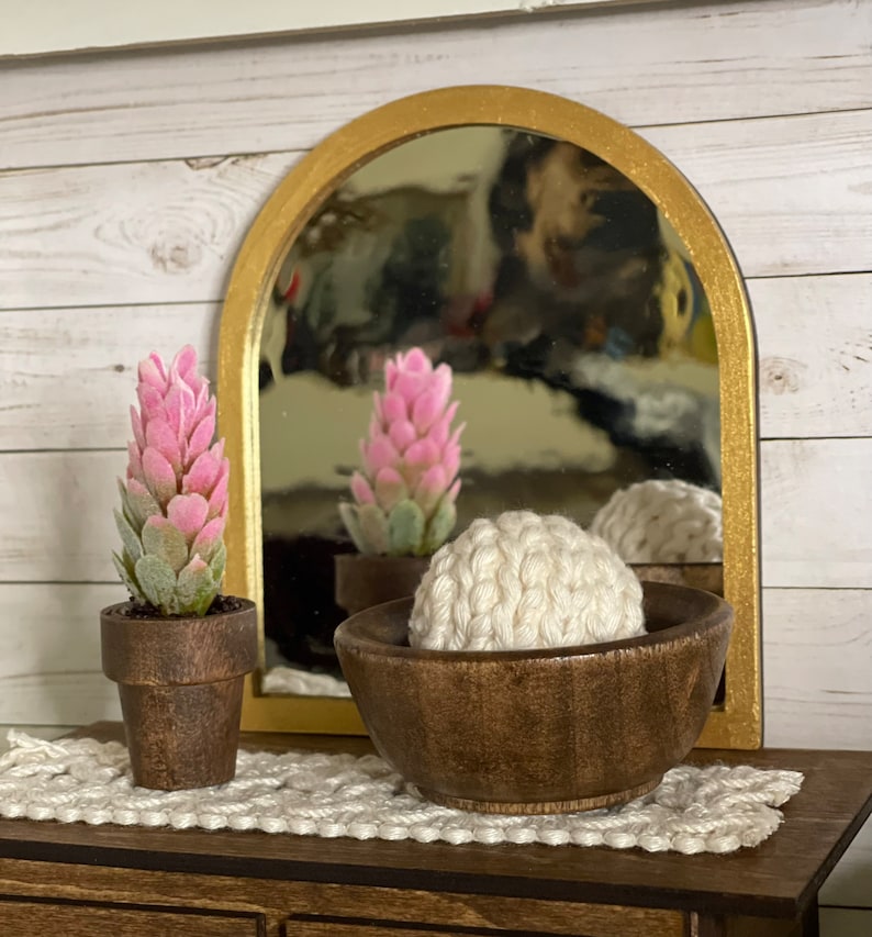 May include: A wooden bowl containing a white crocheted ball, a small potted pink and green plant, and a gold framed arched mirror reflecting a similar plant.