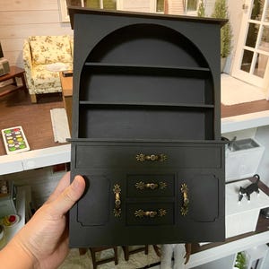 May include: Black wooden hutch with an arched top and three shelves. The bottom section has two doors and three drawers with gold-toned hardware.