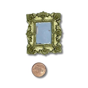 May include: A small, ornate gold-colored mirror with a rectangular reflective surface. The frame features intricate floral and scrollwork designs. A penny is placed below the mirror for size comparison.