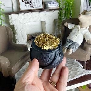 May include: A miniature black cauldron overflowing with gold coins, held in a hand. The scene includes a dollhouse interior with a fireplace, framed artwork, and a stuffed animal in a chair. The overall theme is whimsical and decorative.