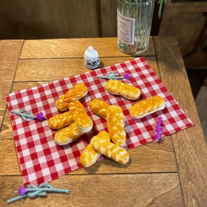 May include: Miniature artisan bread rolls with a golden crust are arranged on a red and white checkered cloth. The scene includes a small sugar shaker, a vase with white flowers, and sprigs of lavender, all set on a wooden table.