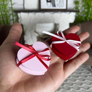 May include: Two heart-shaped boxes, one pink and one red, are held in a hand. Each box is tied with a ribbon bow. The pink box has a red ribbon, and the red box has a white ribbon. A framed black and white picture hangs in the background.