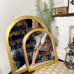 May include: Two gold arched mirrors, one slightly larger than the other, are leaning against a wall. The mirrors are reflecting a doll sitting on a chair.