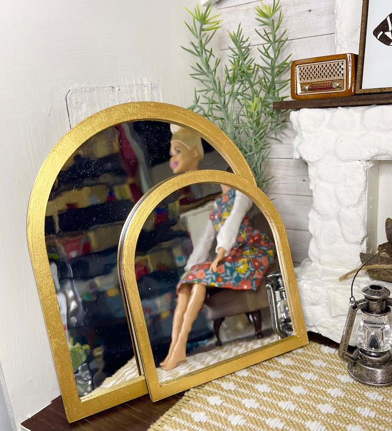 May include: Two gold arched mirrors, one slightly larger than the other, are leaning against a wall. The mirrors are reflecting a doll sitting on a chair.