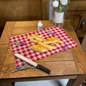 May include: Miniature scene featuring a wooden table with a red and white checkered cloth. Several miniature baguettes are arranged on the cloth, alongside a serrated knife. A small bottle and a vase of white flowers complete the scene.