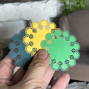 May include: Three paper doilies in shades of green and yellow are held in a hand. Each doily has a scalloped edge with small flower cutouts. The doilies are a craft supply.