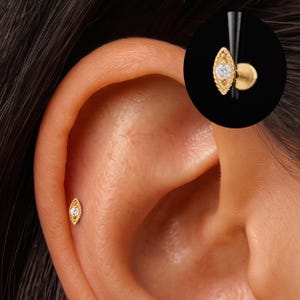 May include: A gold marquise-shaped stud earring featuring a central clear round gemstone. The metal surface has a textured, beaded border design. The earring is shown inserted into the ear cartilage and also as a separate, enlarged view.