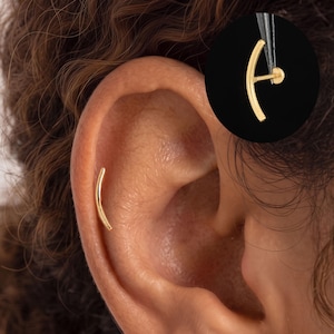 May include: A gold-toned curved bar earring stud featuring a smooth, polished finish. The earring is shown inserted into the outer edge of an ear, with an additional view in a circular inset displaying the post and flat back mechanism.