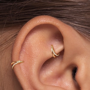 Puede incluir: Primer plano de dos pendientes de aro dorados con pequeñas piedras preciosas transparentes. Un pendiente está en el hélix y el otro en la concha de una oreja. Los pendientes tienen un diseño delicado y curvado, y las piedras preciosas añaden un toque de brillo.