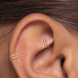 May include: Close-up of an ear adorned with two gold-colored, double-hoop earrings. Each earring is encrusted with small, sparkling stones, creating a delicate and elegant appearance. The earrings are positioned on the ear's cartilage and lobe.