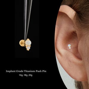 May include: A gold-colored titanium push pin earring with a white opal and two clear crystals. The earring is shown in the ear and held by a tool against a black background. Text on the image reads "Implant Grade Titanium Push Pin 16g. 18g. 20g."