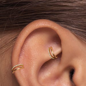 May include: Close-up of an ear with two gold-colored hoop earrings. Each earring has a double band design, with one band adorned with small, clear gemstones. The earrings are set against the skin of the ear.