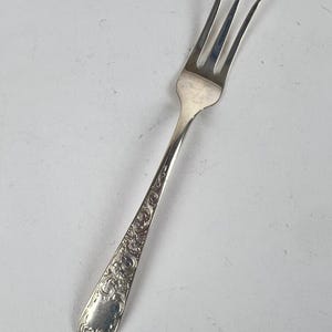 May include: A silver-toned metal cocktail fork featuring three slender tines and an ornate, embossed floral scrollwork pattern along the length of the handle. The handle tapers toward the base, showcasing a detailed, reflective metallic finish.