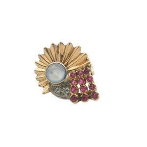 May include: A gold-toned ring with a sunburst design, a central moonstone, and a cluster of small red gemstones. The ring also features small clear stones. The ring is set against a white background.
