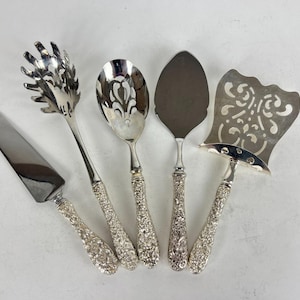 May include: A set of five silver-toned serving utensils featuring ornate, textured handles. The collection includes a flat cake server, a pasta fork with long tines, a slotted spoon, a solid spade-shaped server, and a wide, decorative pie server.