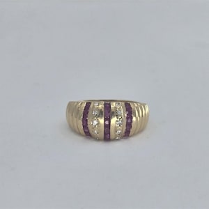 May include: A gold ring with a ribbed design. The ring features alternating vertical bands of small, round, red gemstones and clear, round gemstones. The ring is set against a plain white background.