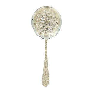 May include: A silver-toned serving spoon featuring a wide, circular bowl with an embossed design of a cluster of grapes and leaves. The handle is long and slender, decorated with a detailed, intricate floral pattern that extends down its length.