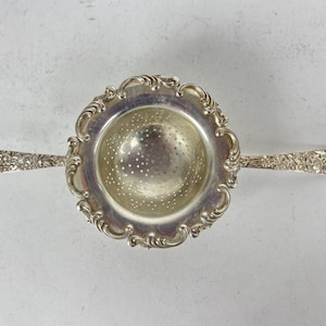 May include: A silver tea strainer with a decorative floral design on the handles and rim. The strainer has a perforated bowl for filtering tea leaves. The handles extend outward from the bowl.