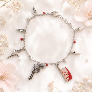 May include: A silver-toned chain charm bracelet featuring multiple metal charms including a revolver, a cleaver with red paint splatters, a four-pointed star, and several conical spikes, accented with small white pearls and tiny red beads.