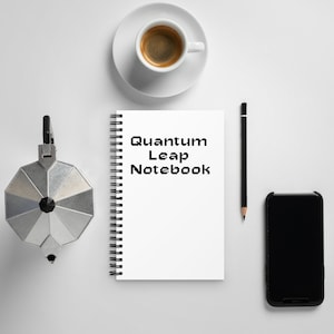 May include: A white notebook with a black spiral binding and the text "Quantum Leap Notebook" printed on the front. The notebook is on a white surface with a coffee cup, a pencil, and a black smartphone.