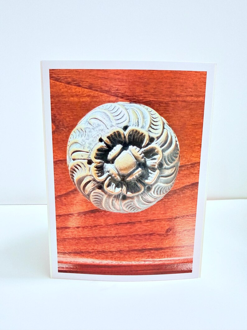 Puede incluir: Un pomo de mueble redondo de metal con acabado en lat&oacute;n antiguo, que presenta un patr&oacute;n floral detallado en relieve con p&eacute;talos en espiral que irradian desde el centro, montado sobre una superficie de madera pulida de color marr&oacute;n rojizo.