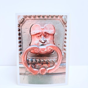Puede incluir: Una tarjeta de felicitaci&oacute;n rectangular con una fotograf&iacute;a de un llamador de puerta de metal marr&oacute;n oscuro. El llamador tiene forma de rostro humano estilizado con una frente prominente y un asa curva y ornamentada que cuelga de la boca.