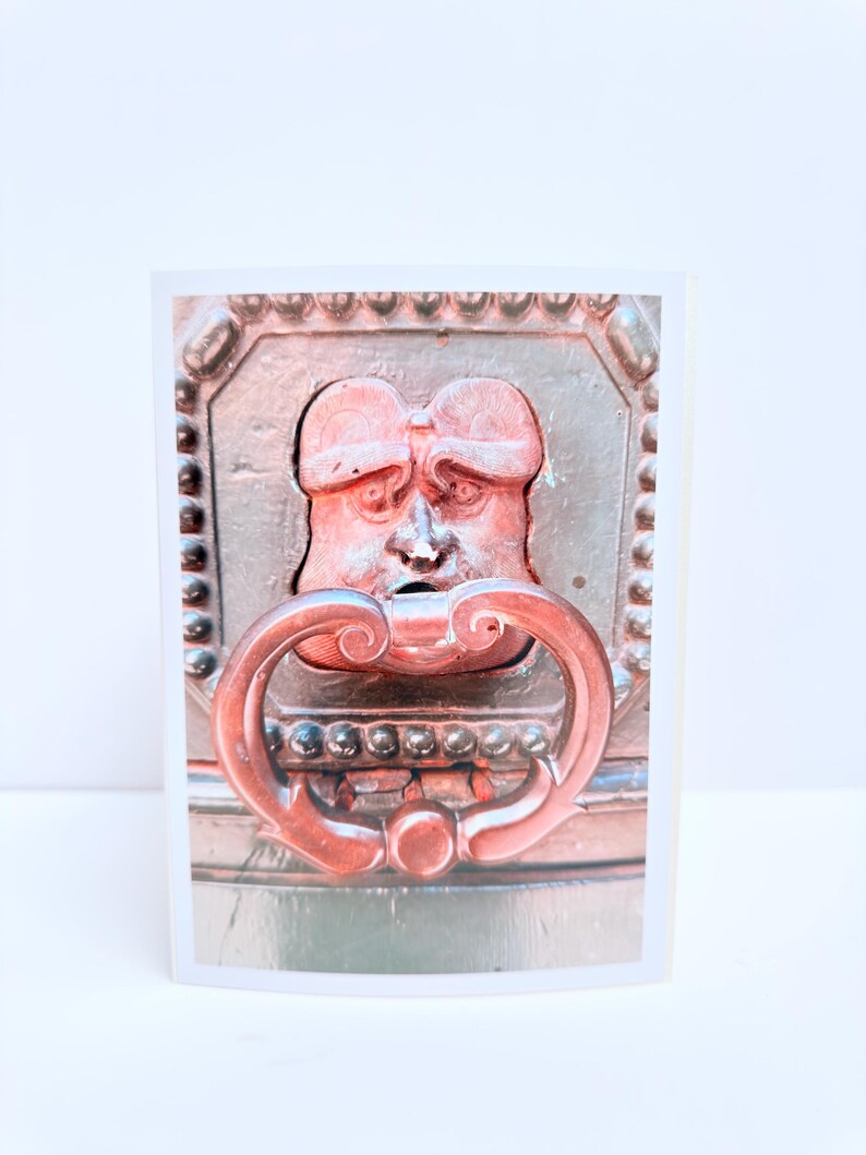 Puede incluir: Una tarjeta de felicitaci&oacute;n rectangular con una fotograf&iacute;a de un llamador de puerta de metal marr&oacute;n oscuro. El llamador tiene forma de rostro humano estilizado con una frente prominente y un asa curva y ornamentada que cuelga de la boca.